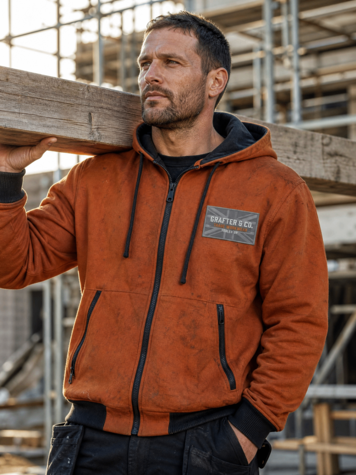 Tradesman on a scaffolded site wearing an orange Grafter & Co. zip hoodie while carrying a timber beam