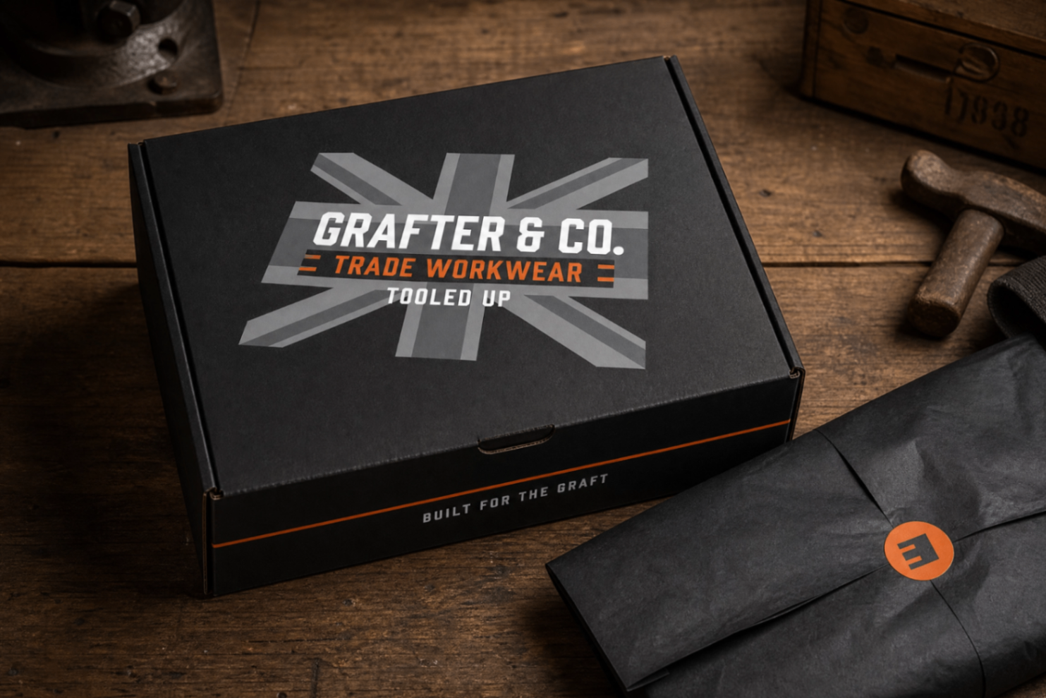 Black Grafter & Co. mailer box with grey union jack print, orange base stripe and 'Built for the graft' tagline on a workbench with tools